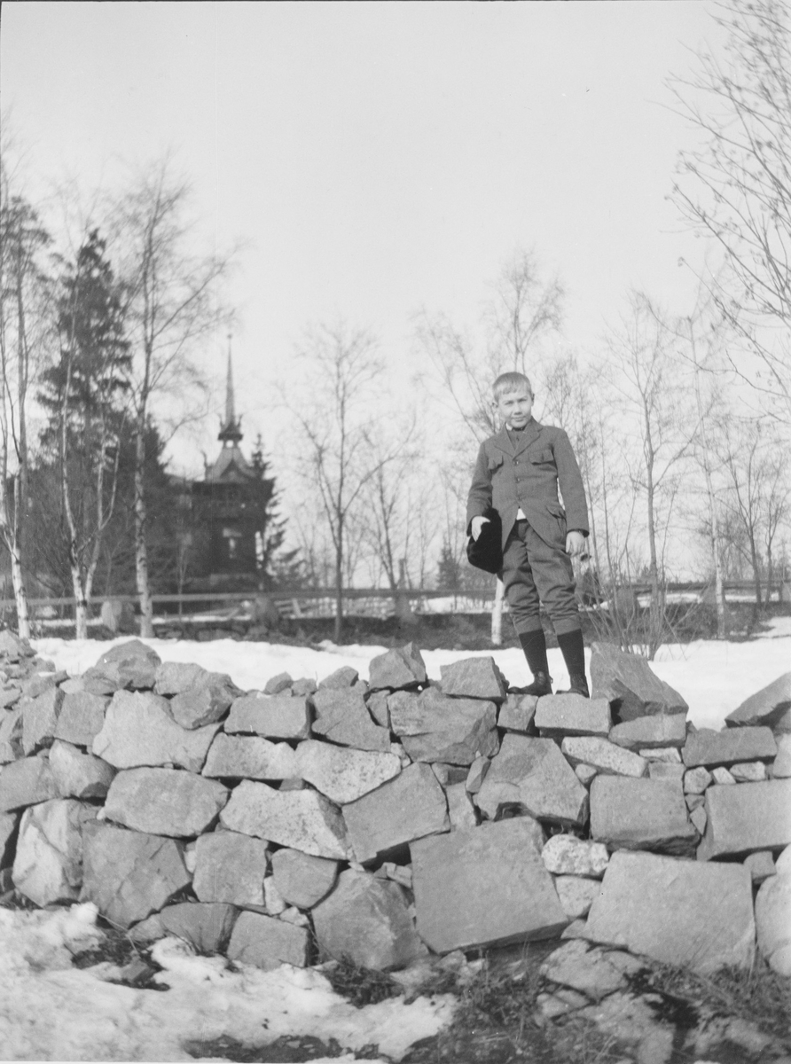 Bildet er kanskje tatt ved Holmenkollen Turisthotel og det er Iacob Ihlen Mathiesen som står på toppen av en tørrmur eller steinmur med luen i hånden.