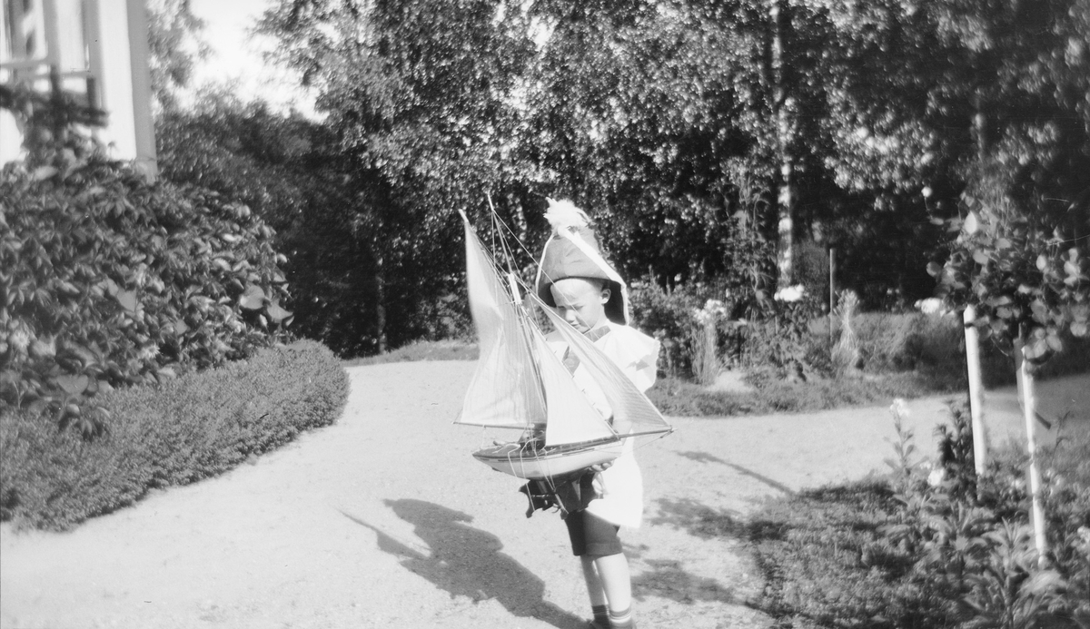 Iacob Ihlen Mathiesen har kledd seg ut som kaptein, har "kapteinshatt" på hodet og holder en liten seilbåt. Han står på gruslagte hagestier, kranset med frodige blomsterbed, utenfor hovedhuset.