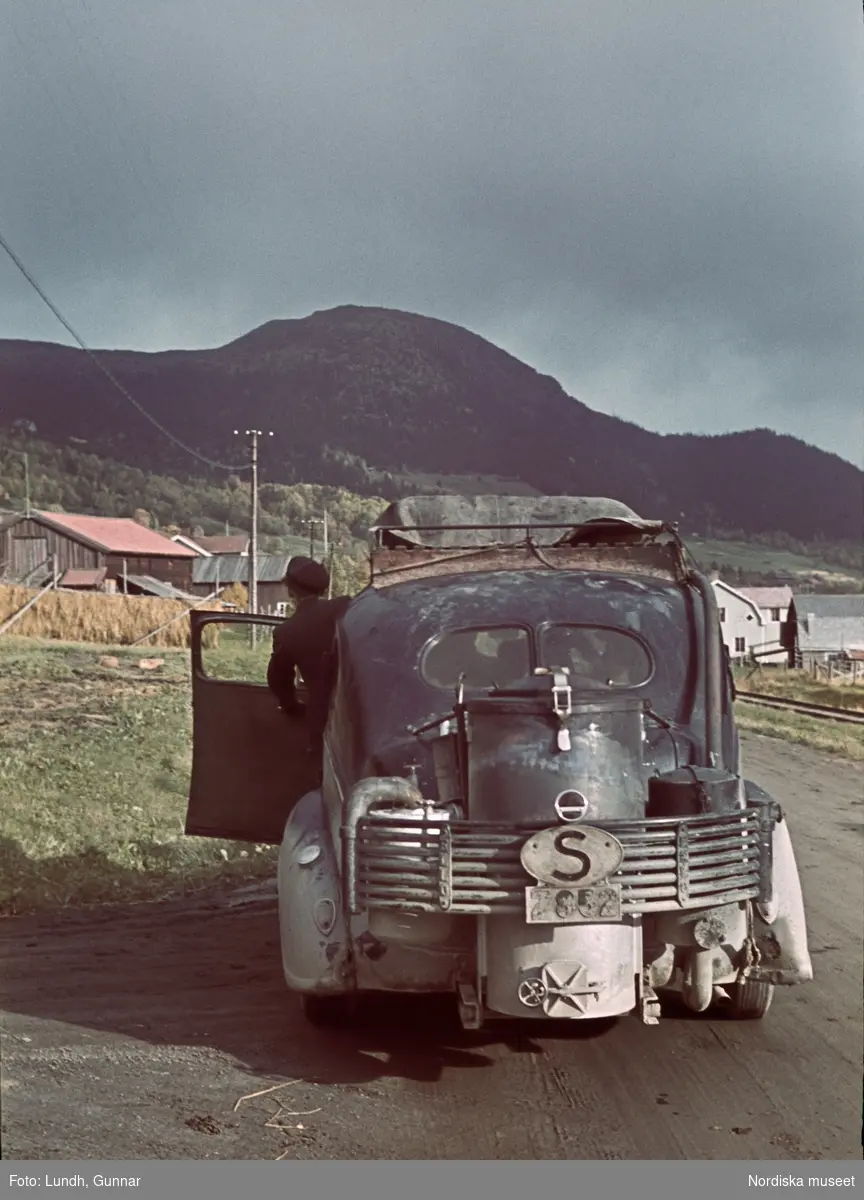 En bil med gengasaggregat har stannat på landsvägen till en fjällby i Åre år 1943. En man i skämmössa och svart kavaj (möjligen uniformsjacka för chaufförer?) kikar ut genom förarplatsens öppna dörr. I bakgrunden fjället Totthummeln. Bilen har länsboksatvsbeteckningen Z för Jämtland.