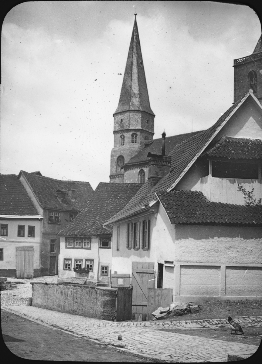 Skioptikonbild med motiv från Hafenmarkt i Münnerstadt med Stadtpfarrkirche St. Maria Magdalena i bakgrunden.
Bilden har förvarats i kartong märkt: Vårresan 1910.Münnerstadt.