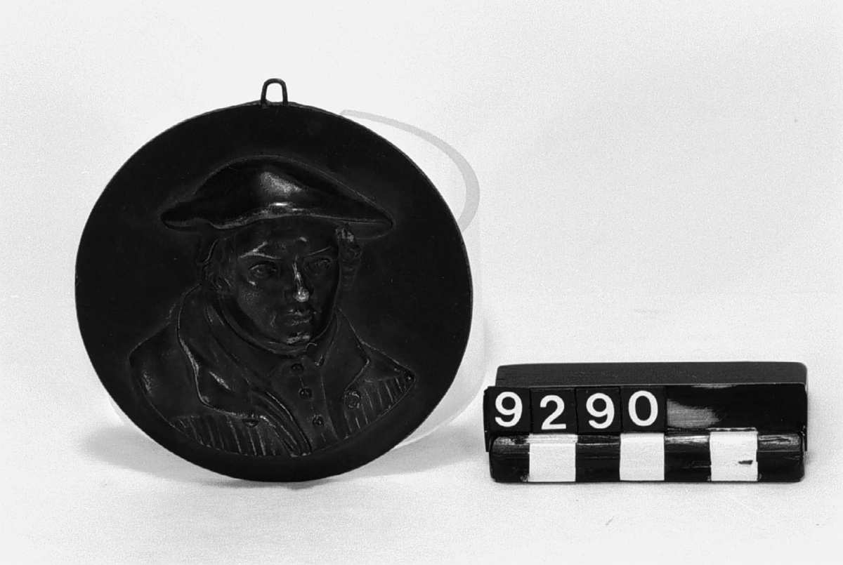 Medaljong av gjutjärn, mansporträtt i hög relief, förmodligen Martin Luther. Märkt på baksidan: "Geiss". Tjocklek: 14 mm.