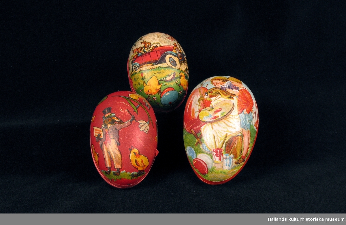 Påskägg av pressad papp i två halvor. Två mångfärgade bilder, på ena halvan drar två kycklingar en kärra med ett ägg på, hönan och tuppen ser på. Den andra halvan visar en hare som, utklädd till konstnär, målar ett ägg, två barn ser på.