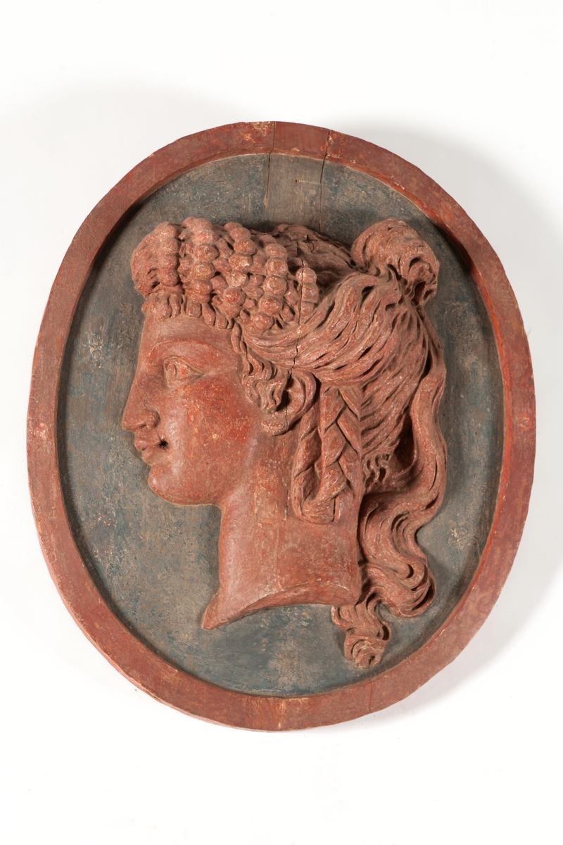 Medaljong, antikiserat kvinnohuvud, enligt uppgift föreställande grevinnan Ehrenswärds huvud. Röd på blågrön botten med oval röd kant.