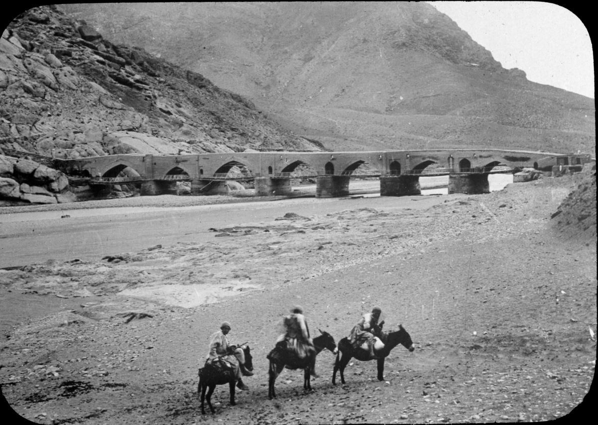 "Bro mellan Rescht - Kazvin." (Rasht-Qazvin) Tre män med packning på åsnor vid flodbädd framför en längre bro. Möjligen bron i Lowshan.