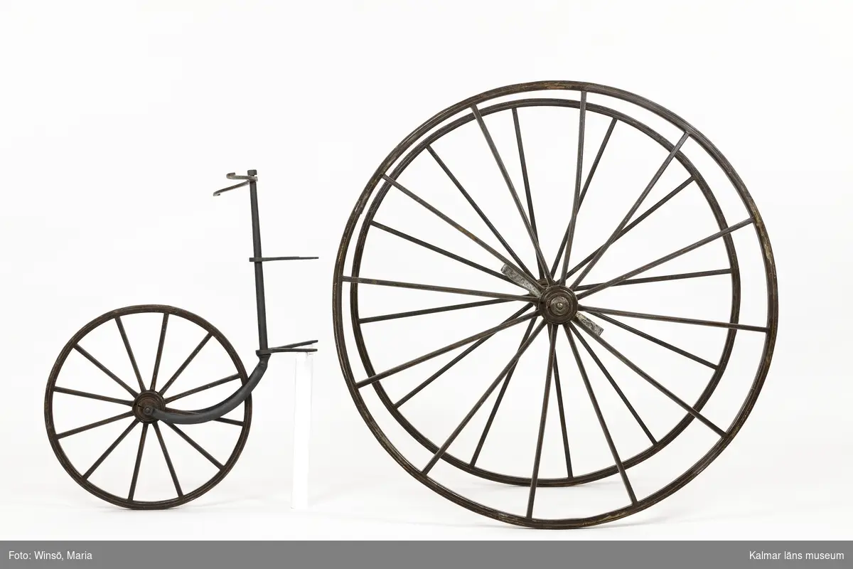 KLM 12224:1-5. Velociped, trehjuling. Fem delar. Av hemgjord typ. Flera delar till ramen saknas. Tre stycken hjul av trä, målade i brunt med gul dekorrand. Hjulen är järnskodda och ett av de stora hjulen har stämplar i järnet: 1868 samt EA inom en cirkel. Mellan de två stora hjulen (:1-:2) sitter en axel av metall (:3) för trampor, trissor, sannolikt av trä, som kunde snurra runt. Det minsta hjulet (:4) har fungerat som fram- eller bakhjul. På hjulet är en del av ramen monterad. Längst upp ett styrstagsliknande tvärstag (:5) med ett litet runt genomgående hål i respektive ände för infästning, i detta fästes sannolikt det som reglerade styrningen. Själva tvärstaget har inte använts som styre.