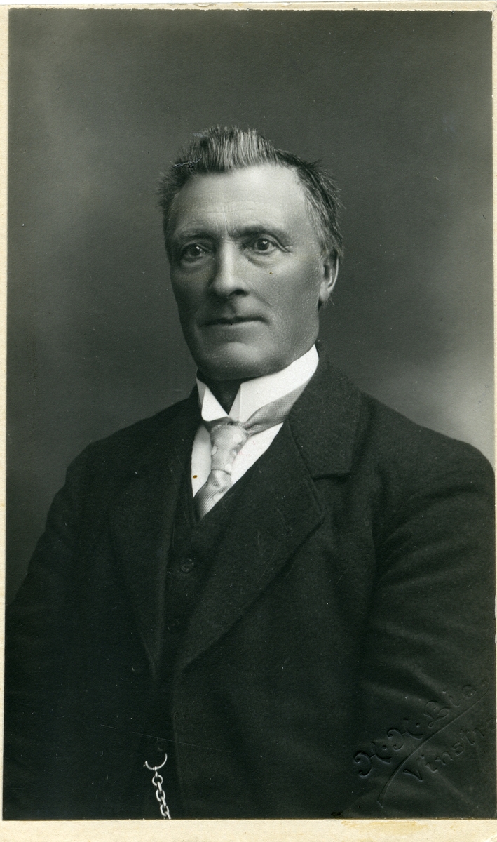 Portrett av en mann sittende foran et lerret iført dress og skjorte med slips.