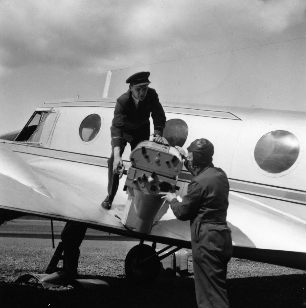 Landskap. Et fly, Avro Anson fra Widerøe, parkert. To personer løfter et avansert fotoapparat inn i flyet.