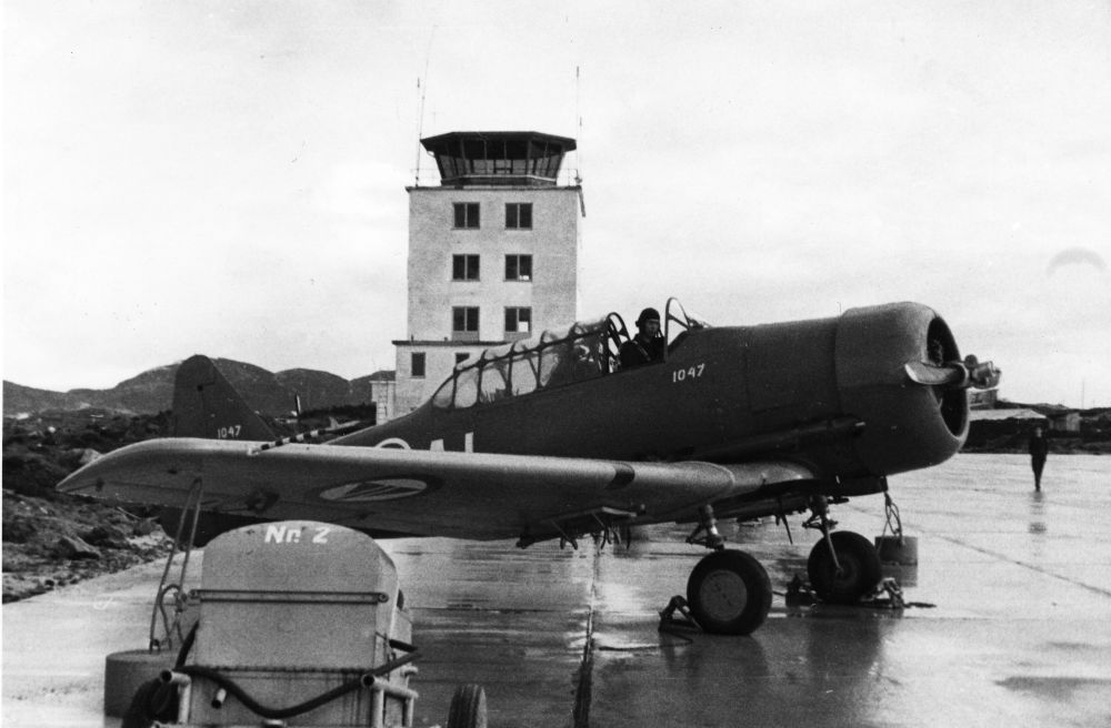 Lufthavn/Flyplass. Bodø. Et militært fly av typen Harvard, parkert foran Bodø kontrolltårn. En person , flyger/pilot, sitter i cockpit.