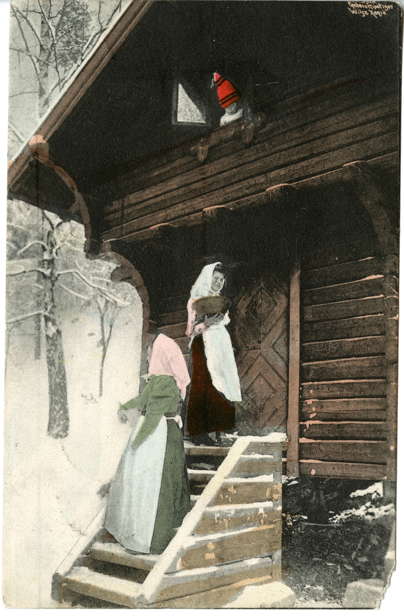 Kolorert postkort. To kvinner på trappa foran stabburet. Liten gutt med nisseluse titter ut fra vinduet.