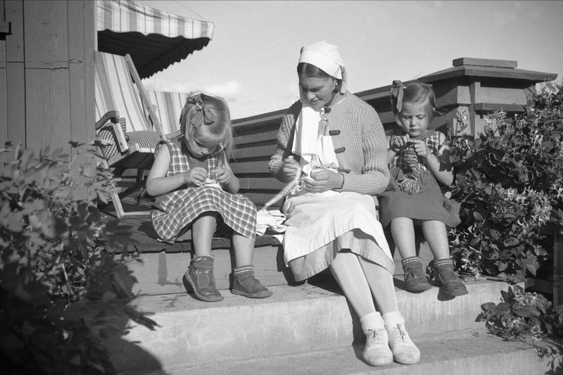 En kvinne og to barn sitter på en tapp og strikker.