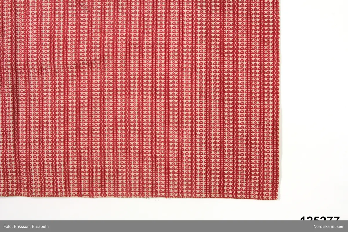 Förkläde i bomull med flerskaftad mönstervävning i rött på vit botten. Täta bårder med små runda bladmönster . 
Mönstervävningen påminner mycket om småmönstrade vävnader moderna under 1820-talet som finns i den tidens vävprovböcker. 
Förklädet har smala handsydda fållar på 3 sidor och är upptill rynkat mot 1,3 cm bred linning av annat randigt förklädestyg .Smalrandiga röda bomullsbandt fastsydda som knytband i linningen.
Anm. I nyskick
Dessa bladförkläden vävdes sannolikt av speciella väverskor.
/Berit Eldvik 2012-03-26