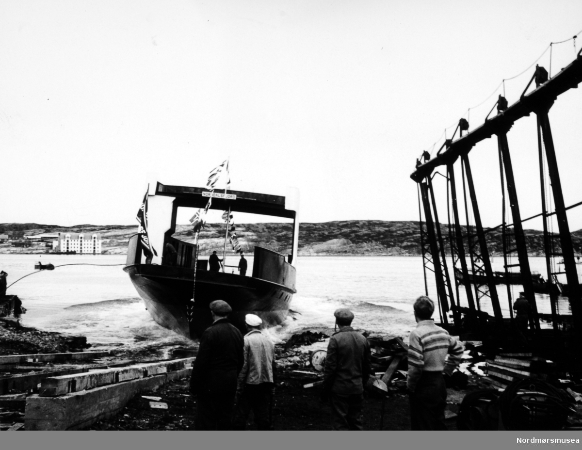 Bildet viser Storviks Mek. Verksted ved sjøsetting av B/F"Norddalsfjord" verkstedets bnr.14 og folkene som bivåner sjøsettingen.

"Norddalsfjord" ble levert til Møre og Romsdal Fylkesbåtar 15. mars 1961 og hadde følgende hoveddimensjoner: L 31,20 m x B 8,55 m x D 3,35 m og hadde en tonnasje på 159 bruttoregistertonn. Fremdriftsmaskineriet består av 3 Volvo Penta turboladede dieselmotorer type TMD96 på til sammen 420 hk som via kilremdrift var koblet til et felles gir og propellaksel med vribar propell, slik at hver enkelt av motorene kunne kjøres separat. Fergen hadde 2 Bolinders vekselstrømsaggregater type 1052MG på 23 hk hver tilkoblet en generator på 17 kW. Fergen var utstyrt med elektrohydraulisk styremaskin.

Fergen har plass til 18 personbiler og har sertifikat for 160 passasjerer. Forut er det innredet 6 lugarer for offiserer og restaurantpersonale og akterut en mannskapslugar for 4 personer og toppfarten er 11,4 knop og marsjfarten 10,5 knop.

Ferga er verkstedets første nybygg etter B/F"Trygge" som ble levert i 1938.

Til høyre ses patentslippvogna klinkbygd i stål 1918, den største slippen mellom Bergen og Trondheim, og slepebåten "Ole Wirum" til Heinsa og dykkerbåten til A/S Anlegg i Trondheim som holder på med reparasjon av slippbanen ute i sjøen. 

Personene foran i bildet er fra venstre: snekker Torvald Amundsen, snekker Johannes Berget, snekker John Betten og sistemann er ukjent. Om bord ses til venstre sjauerformann Carl Clausen og til høyre sjauer John Sund.

I bakgrunnen ses Skorpa med Visnesbrygga og verkstedets fortøyningsbøye, "Storbøya", ute på sundet.

Bildet er fra 1960. Kilde/tekst: Peter Storvik. Fra Nordmøre museums fotosamlinger.
