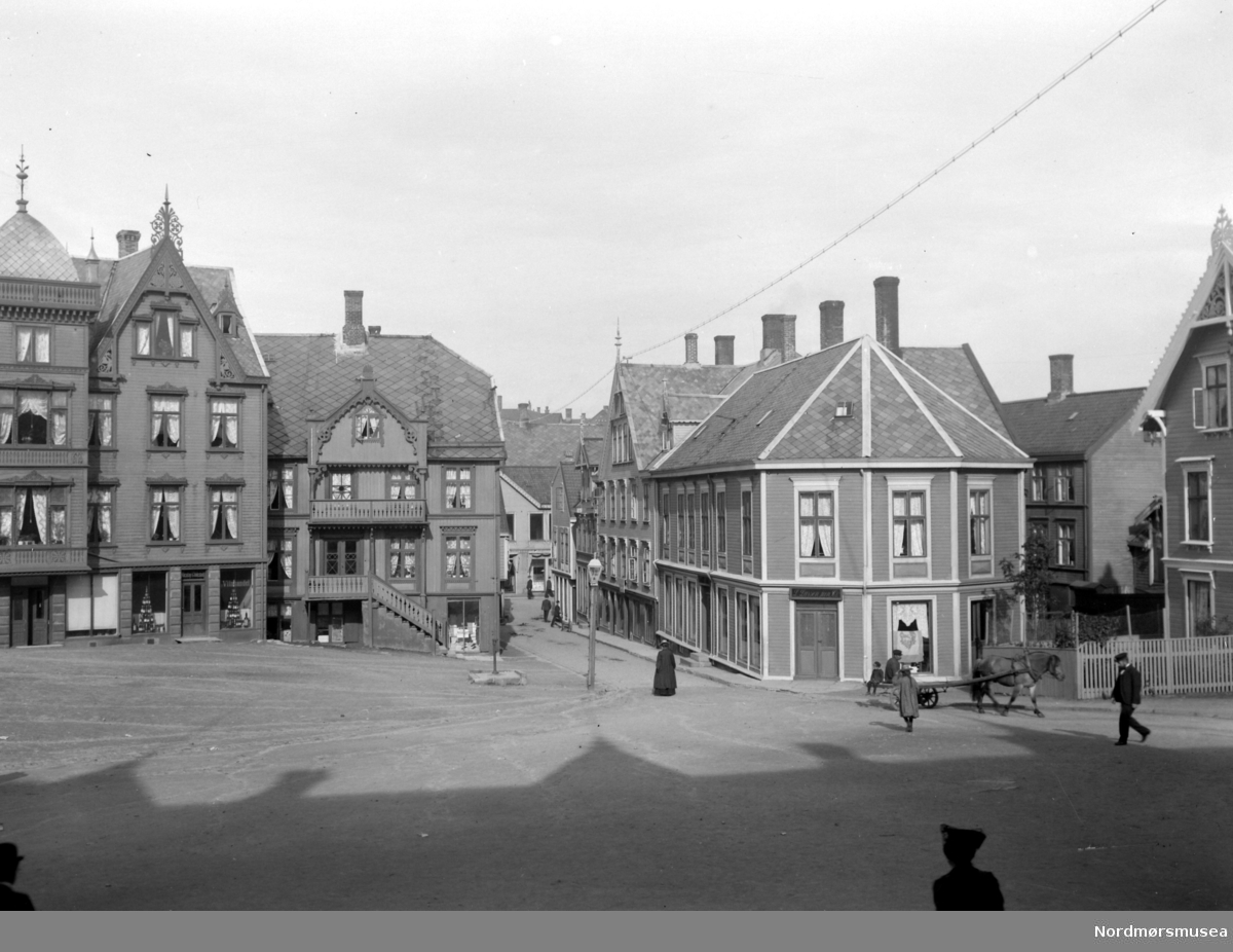 På bildet ser vi blant annet Larsengården til høyre (bygget til høyre med skilt over døren, Parcell M. Nr. 75). Lars Larsen, som eide bygget, kjøpte huset 9. august 1873, og solgte det senere til Fosna Mållag 1. september 1909, som rev bygget fra 1797, og reiste et nytt bygg på tomten. Her hadde Fosna Mållag "Kaffistova" hus fra 1910 til de senere solgte bygget den 1. oktober 1930 til Georg Sverdrup. Bygget brente senere ned under bybrannen 28. april 1940, da tyskerne bombet byen. Fra Nordmøre Museum si fotosamling.
Kilde: Sverdrups bokhandel, 100 år. 1874 - 1974, side 51 og 52.