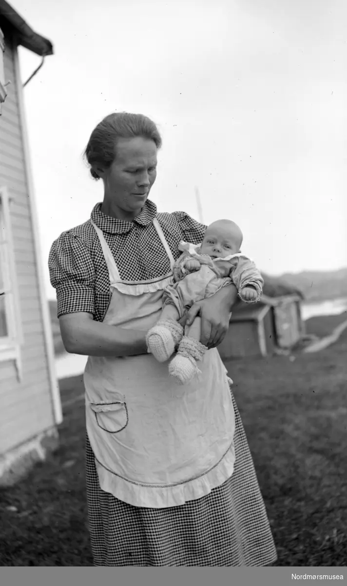 Foto av en kvinne med et barn på armen. Fotografert trolig på Langøya på Vevang i Hustadvika kommune. Smørholmen i bakgrunnen. Fra Nordmøre Museum si fotosamling