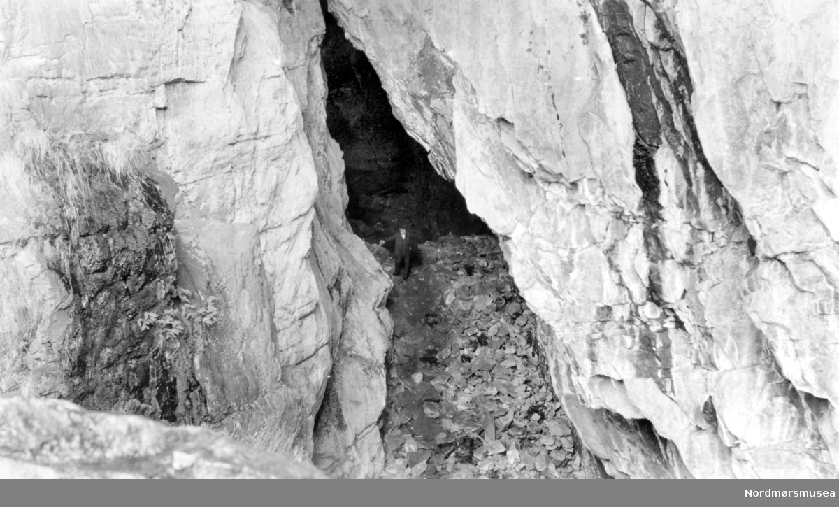  På bildet ser vi Bremsneshula, med inngangen øverst fra steinrøysen. På bildet ser vi en mann som står på toppen av steinrøysen i huleportalen. Fotograf er trolig Georg Sverdrup, og datering er sannsynligvis fra perioden 1930 til 1939. Fra Nordmøre Museums fotosamlinger.
