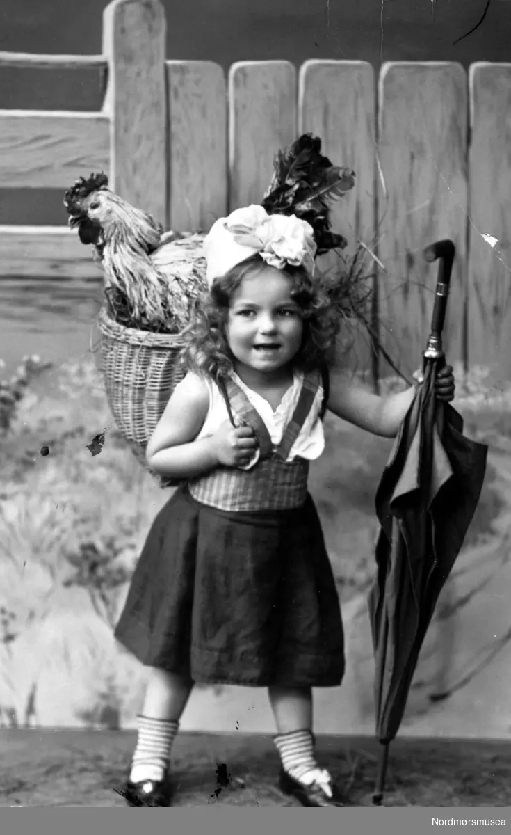 Arrangert postkort, hvor vi ser en ung pike med en paraply og flettekurv på ryggen med to store haner i. Postkortet er antageligvis fra tiden rundt 1910 til 1915. Fra Nordmøre Museums fotosamlinger. Reg: EFR
