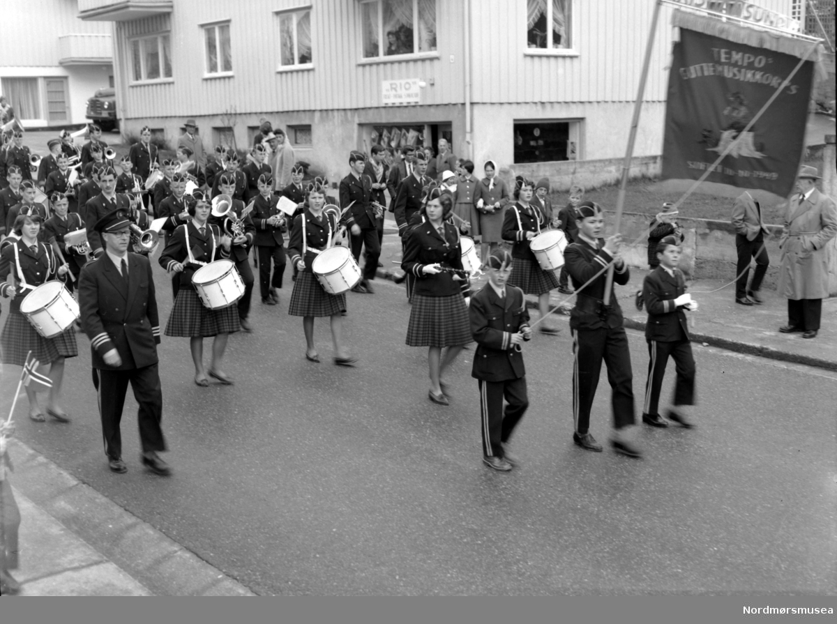 
Tempo guttemusikk med jenter på tromme, marsjerer Hans Neeraas gate, forbi Festiviteten og Rio godeributikk. Fra Nordmøre Museums fotosamlinger.



