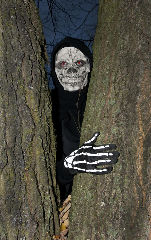 Person utkledde som Døden, med dødnigemaske og skjeletthånd, titter fram mellom to trær.