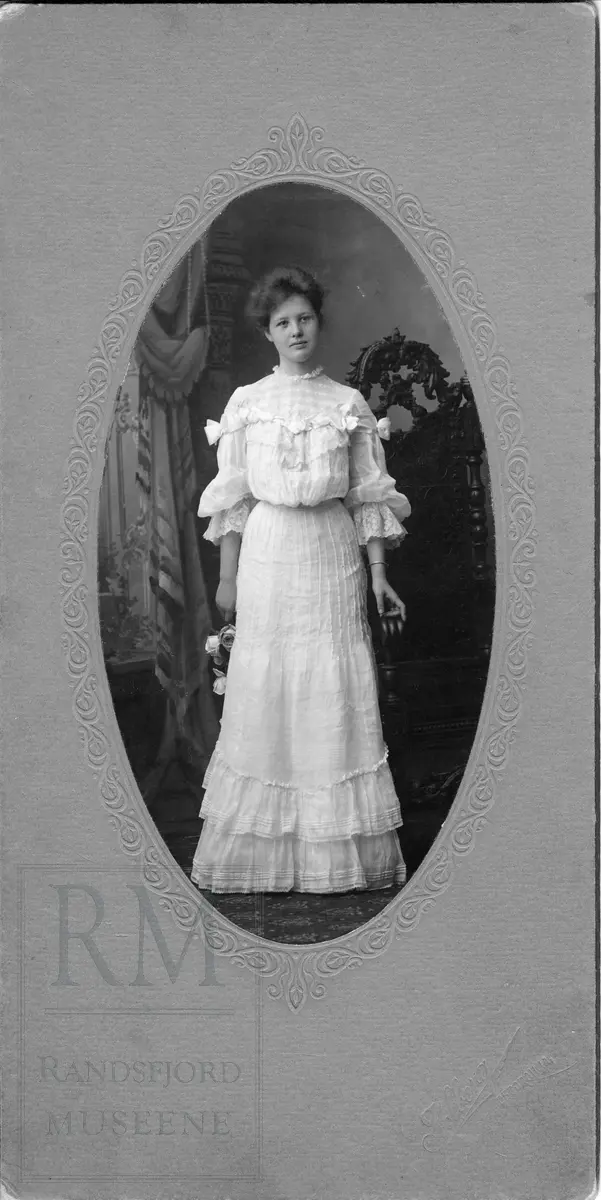 Studiofoto av kvinne i helfigur, i lys kjole, stående ved en rikt dekorert stol og holder en rosebukett i hånden.