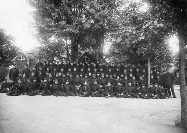Gruppfoto av en pluton meniga med sina officerare ( i mitten och vid sidorna) framför några av de egenartade hyddorna på Kronobergs hed.