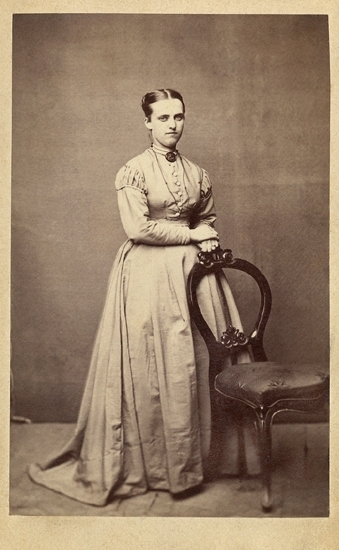 Porträtt (helfigur, halvprofil) av en okänd kvinna i ljus klänning. Vid halsgropen syns en brosch och hon har en kedja runt halsen. 
Hon står snett bakom en stol i nyrokoko.