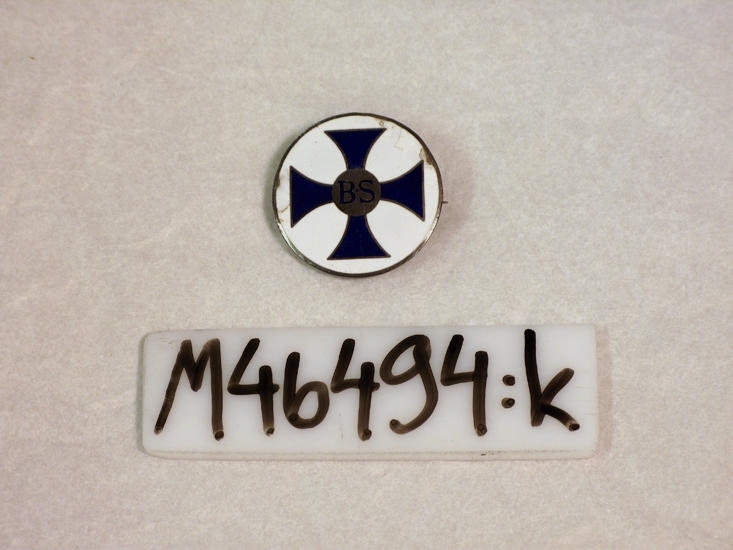 Sjuksköterskeemblem i vit och blå emalj.
Malteserkorset  i blått med silverplatta i mitten och bokstäverna B S ("Barnmorska Stockholm").
Baksidan av silver med stämplar C D S samt V 8- 1947 samt ingraverat i skrivstil; I L (Ingegerd Lander).
Broschen tillhör barnmorskeuniformen M 46494 och är monterad på klänningens krage.


Inskriven i huvudbok  1983
Funktion: Sjuksköterskeemblem