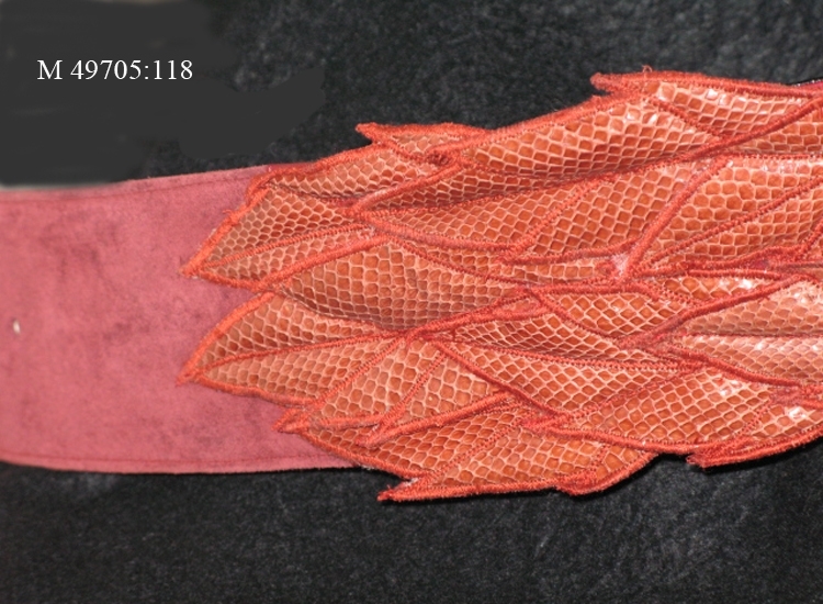 Skärp i plommonfärgad mocka med svart insida av läder.
Mitt fram ett tegelrött 540 mm långt bladformat mönster i relief, av ormskinn.
Märkt inuti på mitten; "Leila Lerkin"


Inskrivet i huvudbok 2010-01-18