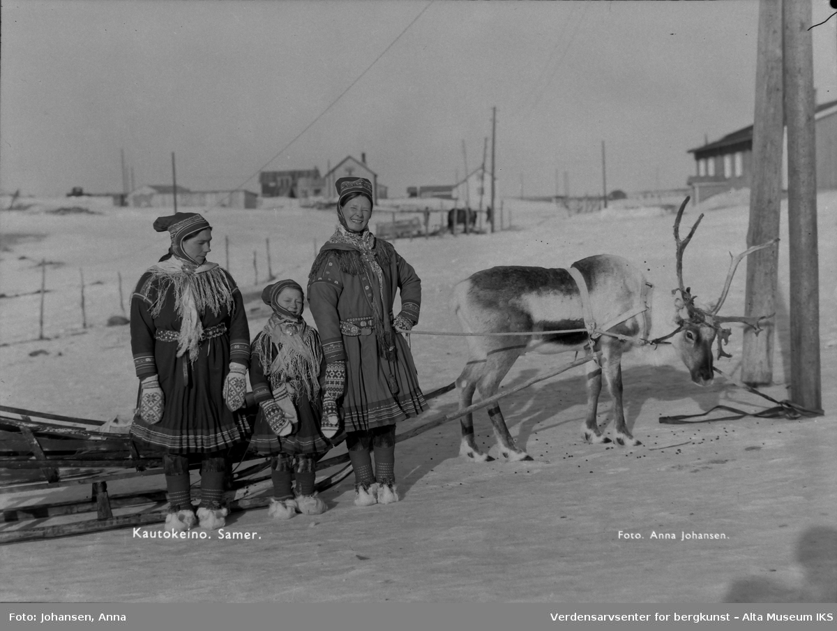 To samiske kvinner og ett barn står foran et reinsdyr med slede. Vinter.