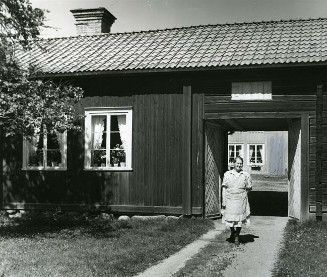 En kvinna iförd förkläde och klänning poserar framför ett portlider vid gården Olsvens.