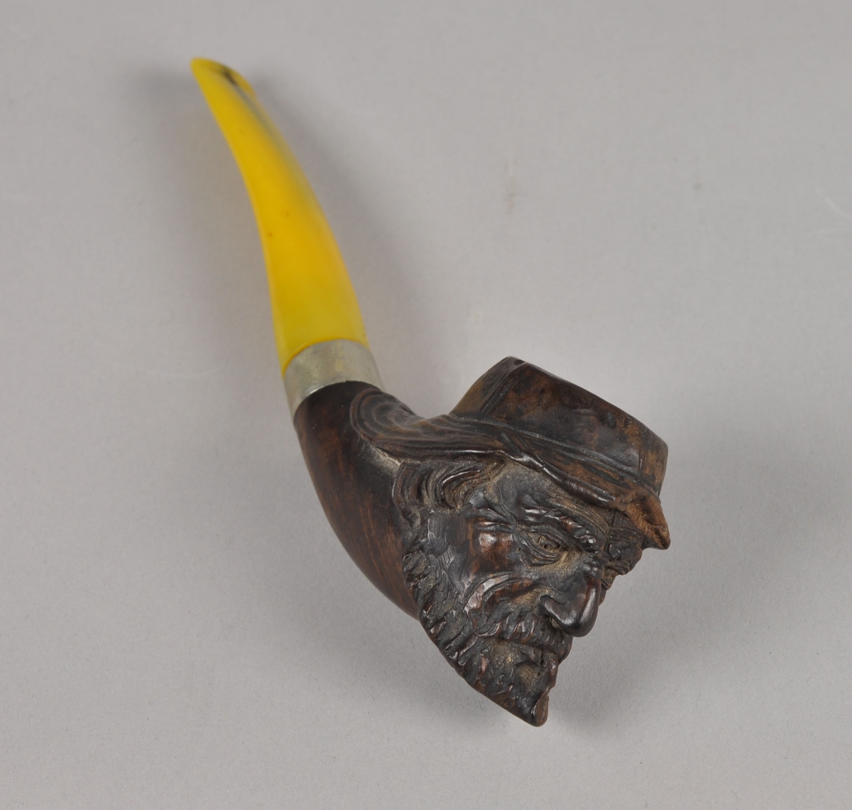 Svakt buet pipe med figurskåret hode av "Fiskeren". Hodet er av tre og munnstykket av kunstig rav. Holk av metall.