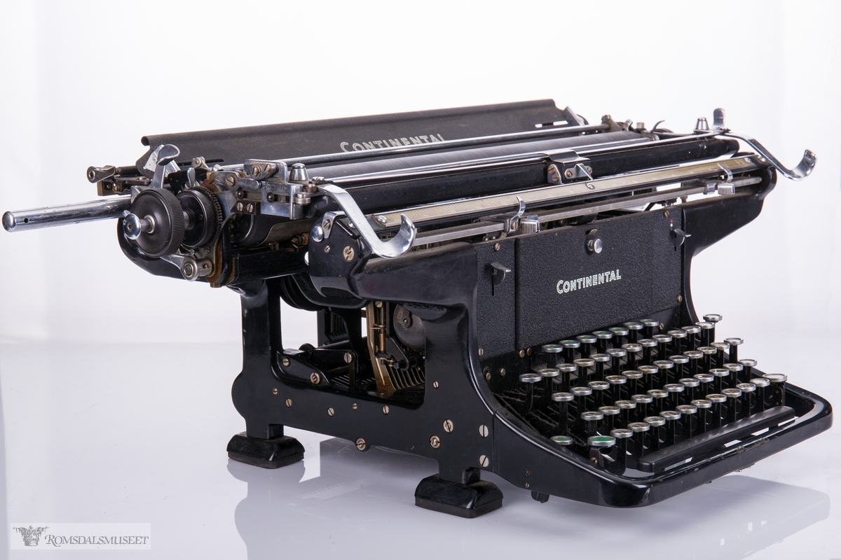 Rektangulær, mekanisk skrivemaskin på fire bein med en bred overdel som går langt utover maskinens grunnform. 
56 tangenter.
