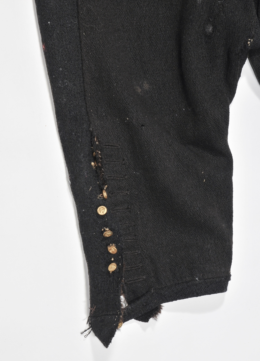Knebukse i svart kypertvove ulltøy med ein bise saum på kvart framstykke.  Det same har saumen midt framme. Linning ved kneet med knapp og knappehol.