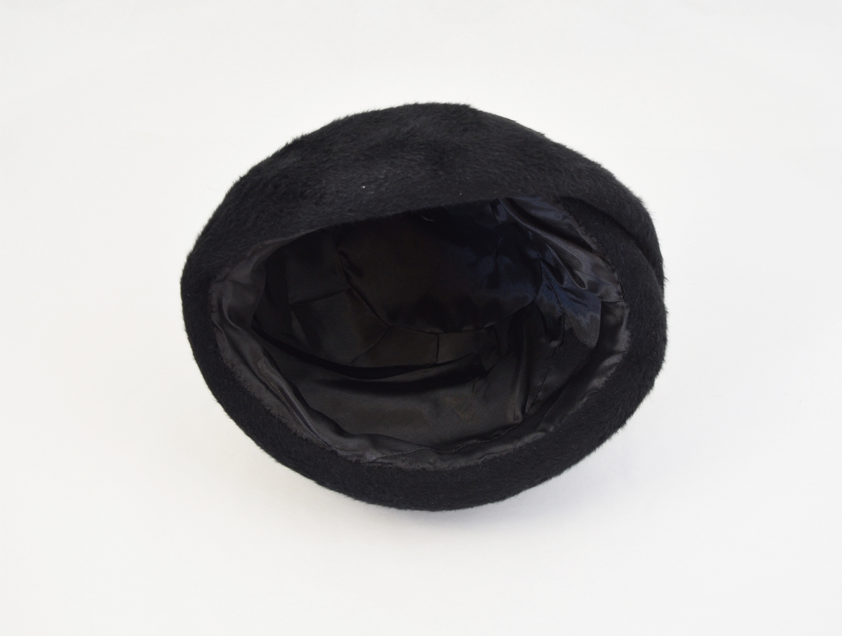 Svart børstet filt. Rund pull, dype lagte folder utgjør hattens fasong. Svart silkefor.