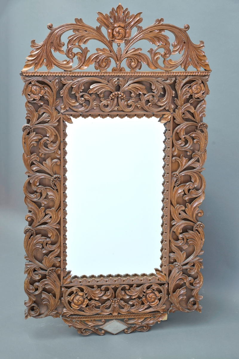 Et firkantet speil i treramme. Rammen er rikt dekorert med utskjæringer i form av blomster og blader.