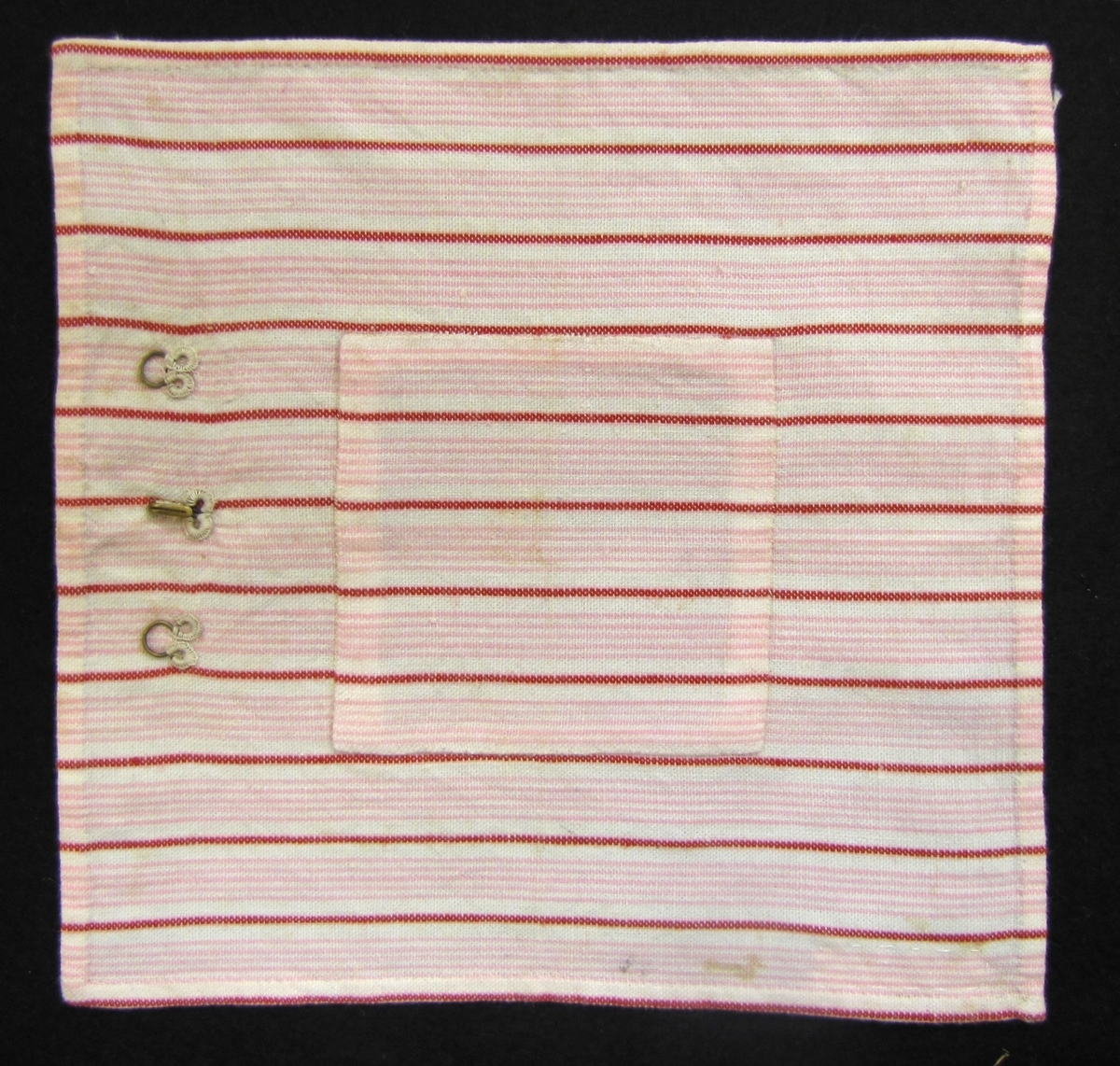 Prov på lappning av lapp på textil. Tygstycke,  randigt i vitt, rött och rosa med en fastsydd hake och två  hyskor. 17 x 18 cm.
Edit Tapper var mor till givarna.