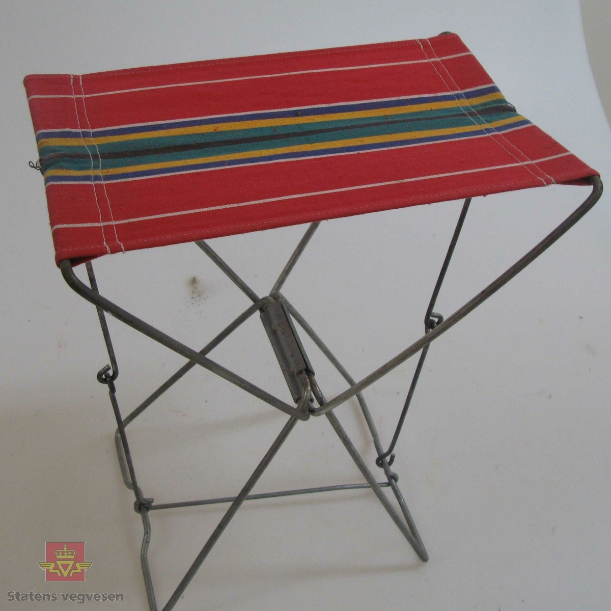 Enkel campingstol med rammeverk av stål og trekk av tekstil. Trekket er flerfarget med striper, hovedfarge er rød. Stolen kan pakkes helt ihop når den ikke er i bruk.
