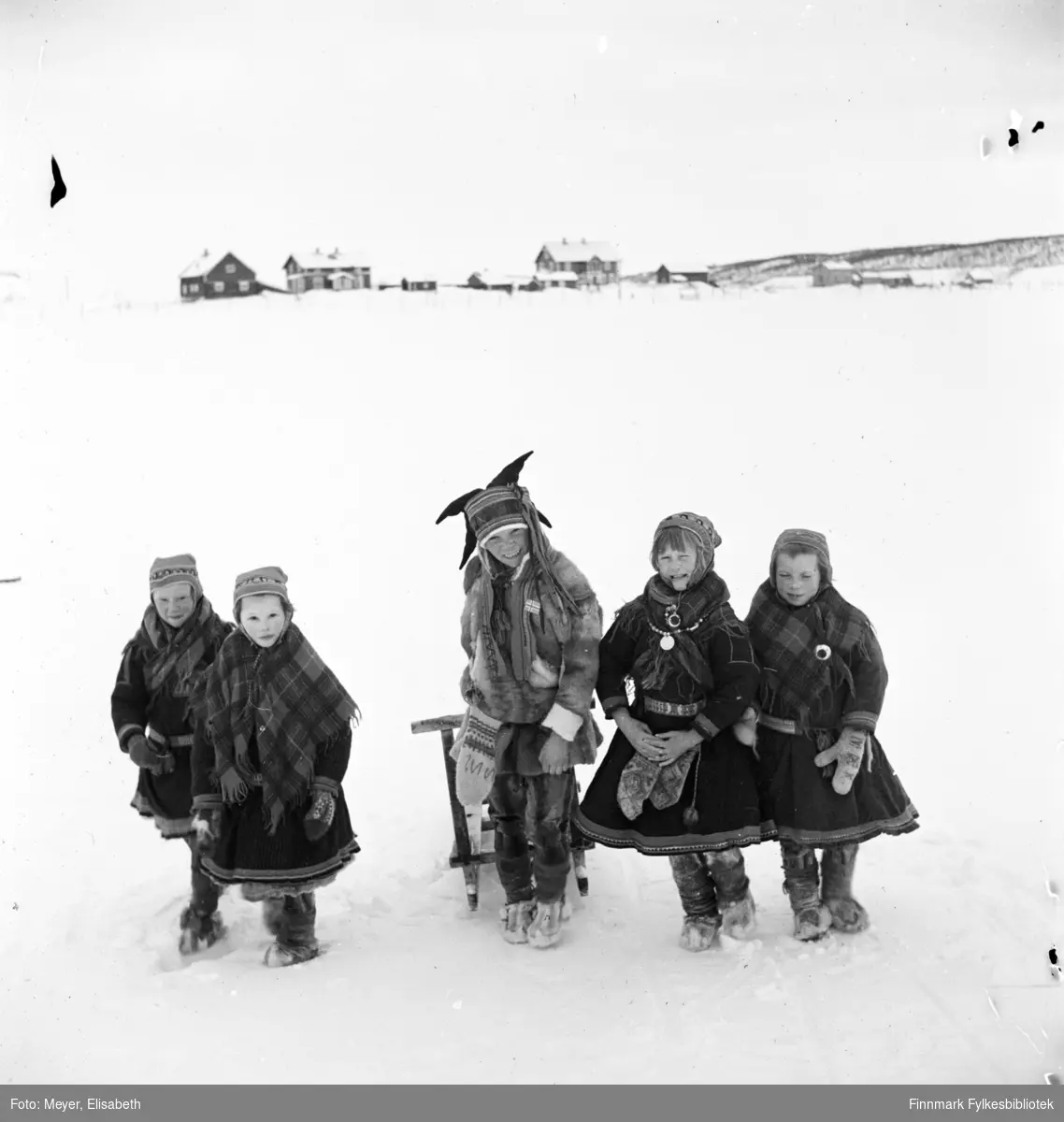 Skolebarn. Fire jenter flankerer en gutt med spark. Alle kledd i samiske kofter og gutten i samisk pesk. Fotografert av Elisabeth Meyer i Kautokeino ved påsketider 1940. I bakgrunnen Prestegården og Lensmannsgården i Kautokeino.