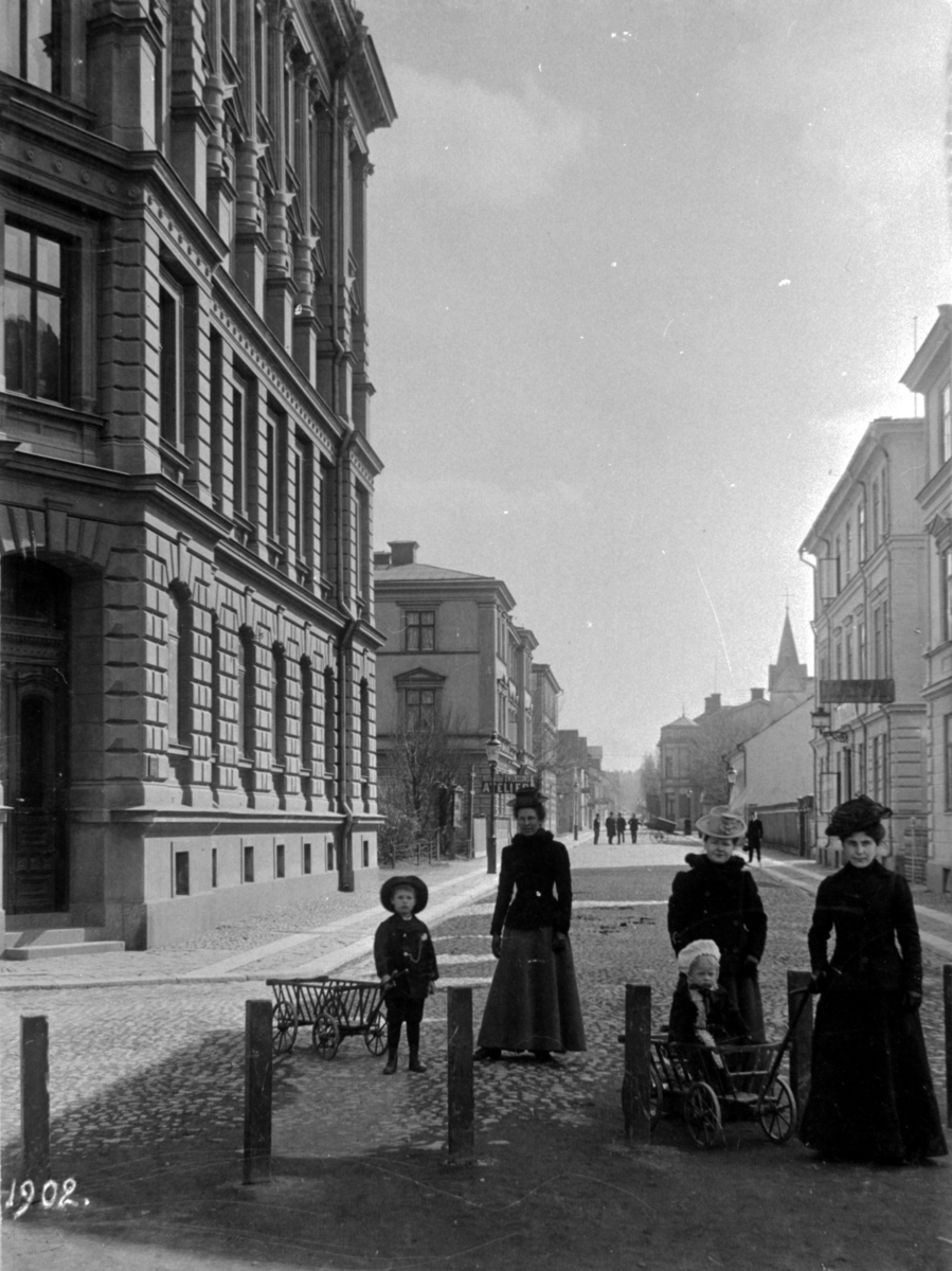 Repslagaregatan söderut från Järnvägsparken.
Tre kvinnor med två barn och två skrindor.
Byggmästare J A Johansson uppförde byggnaden till vänster 1886-88. Arkitekt Janne Lundin, som även gjorde arbets- och detaljritningarna. Huset är typsiskt för 1880-talet. Fasaden ser ut som om det är av sten men är av puts. Ursprungligen fanns det två lägenheter på varje plan, dvs 8 i hela huset. På 1930-talet delades de upp så att huset innehåll 15 lägenheter. Några har på senare tid slagits samman igen.