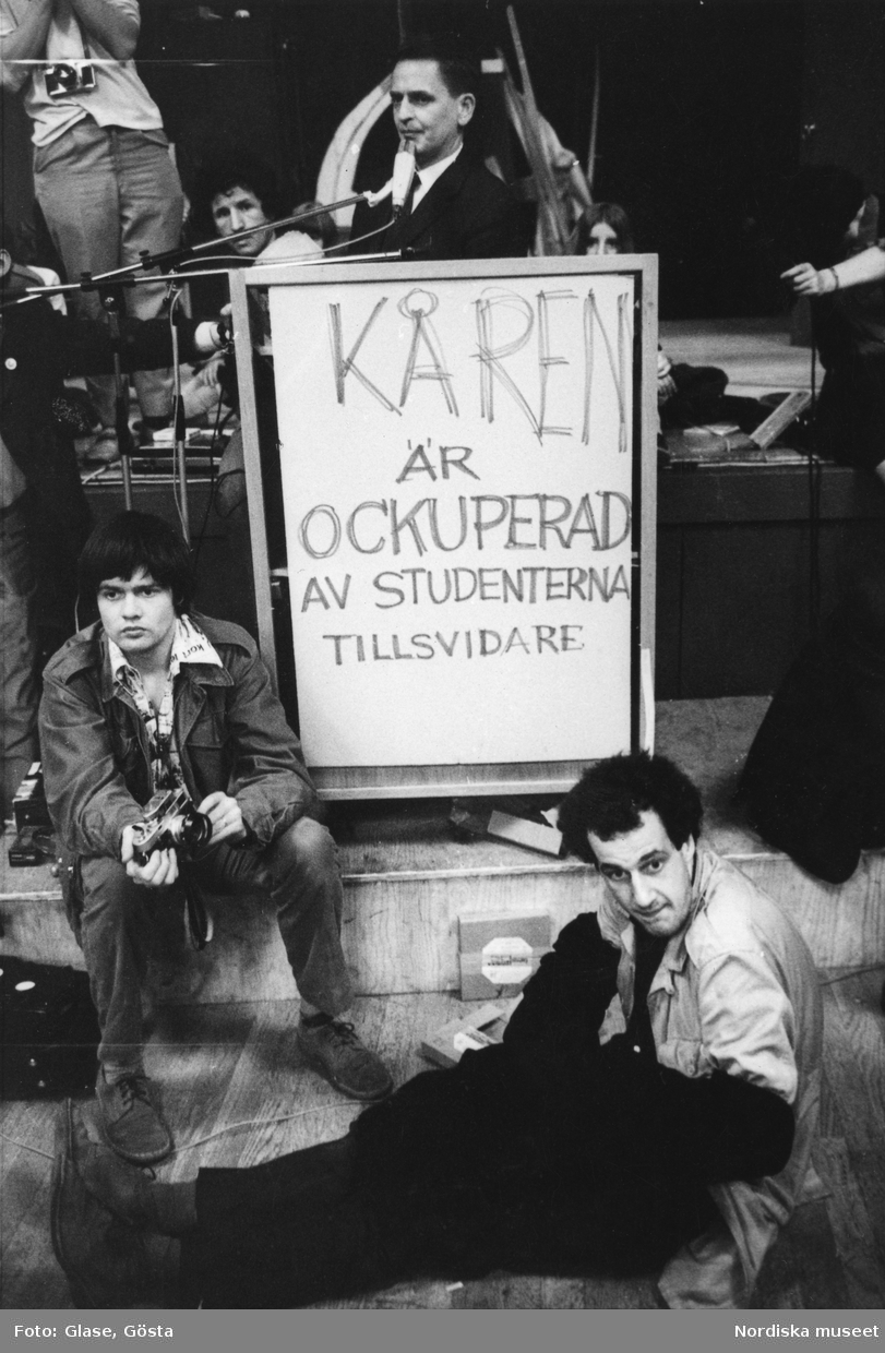 Kårhusockupationen vid Stockholms universitet i slutet av maj 1968. I talarstolen på podiet står dåvarande utbildningsminister Olof Palme. Ett plakat bär texten KÅREN ÄR OCKUPERAD AV STUDENTERNA TILLSVIDARE. Framför sitter två unga män; till vänster konstnären Carl Johan de Geer som fotograferade på uppdrag av tidningen Se, till höger Staffan Lamm filmare åt SVT.