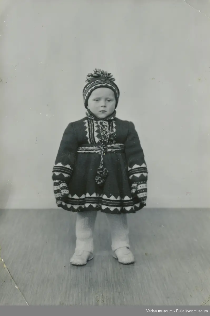 Bjørg Feldt som barn, oppstilt med hjemmestrikket drakt bestående av kofte, lue og votter.