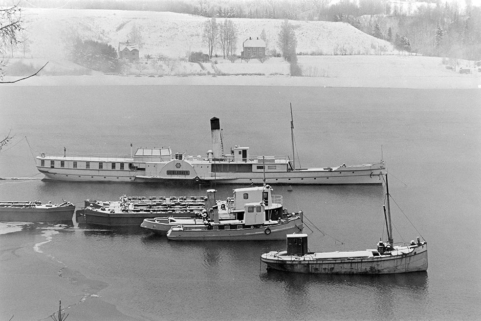 Mjøsbåter, vinteropplag, Minnesund, Minne, Skibladner, tømmerbåter, Fra bakerst i bildet: Skibladner (bygget 1856), 3 mudderlektere (de eldste bygget 1930), tømmertrekkerne Sævat (bygget 1920) og Winnie (til Mjøsa 1949), lastebåten Svalen II (bygget 1923).