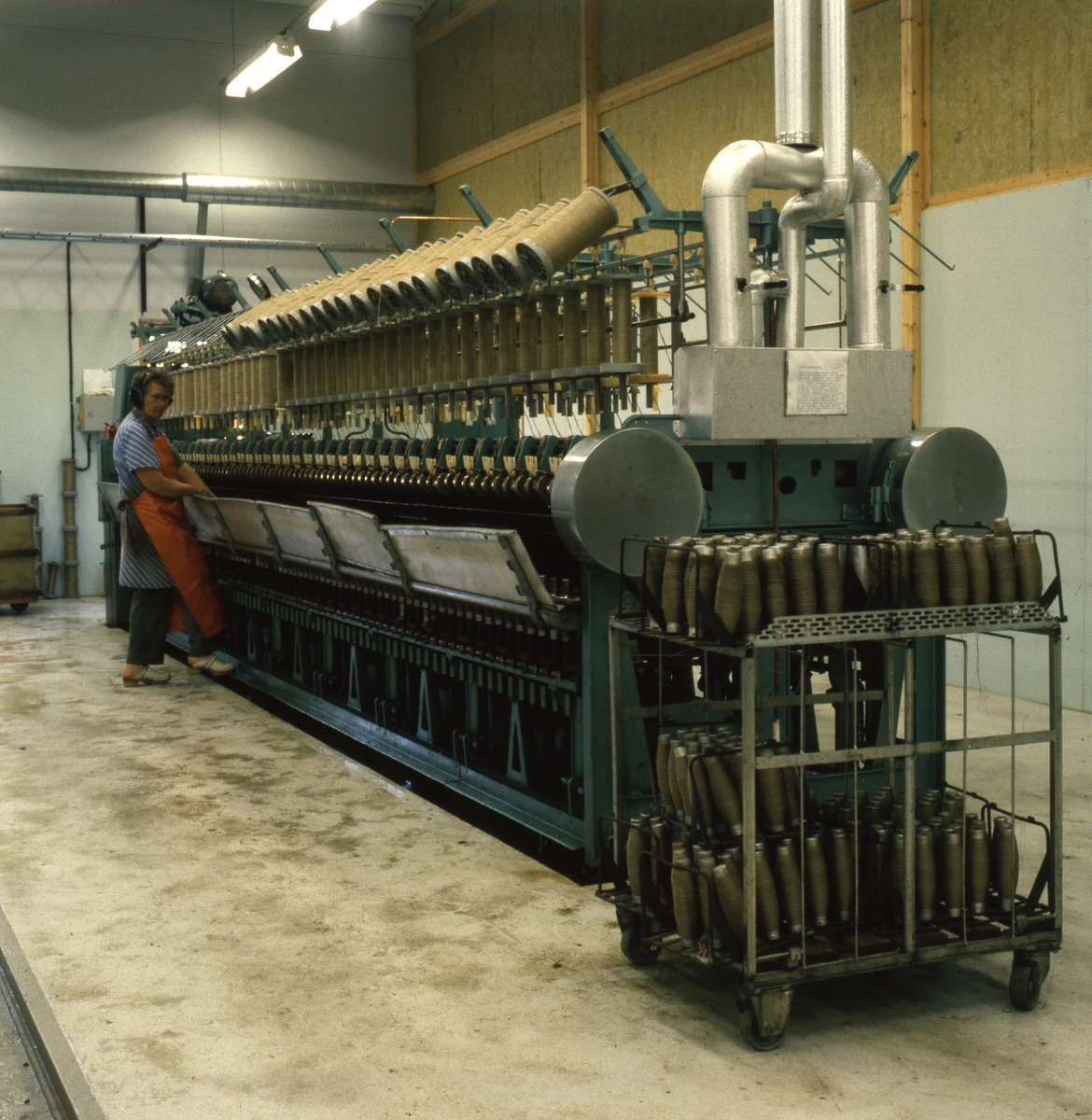 En kvinna står och sköter en stor maskin med bobiner med lintråd på i en industrilokal.