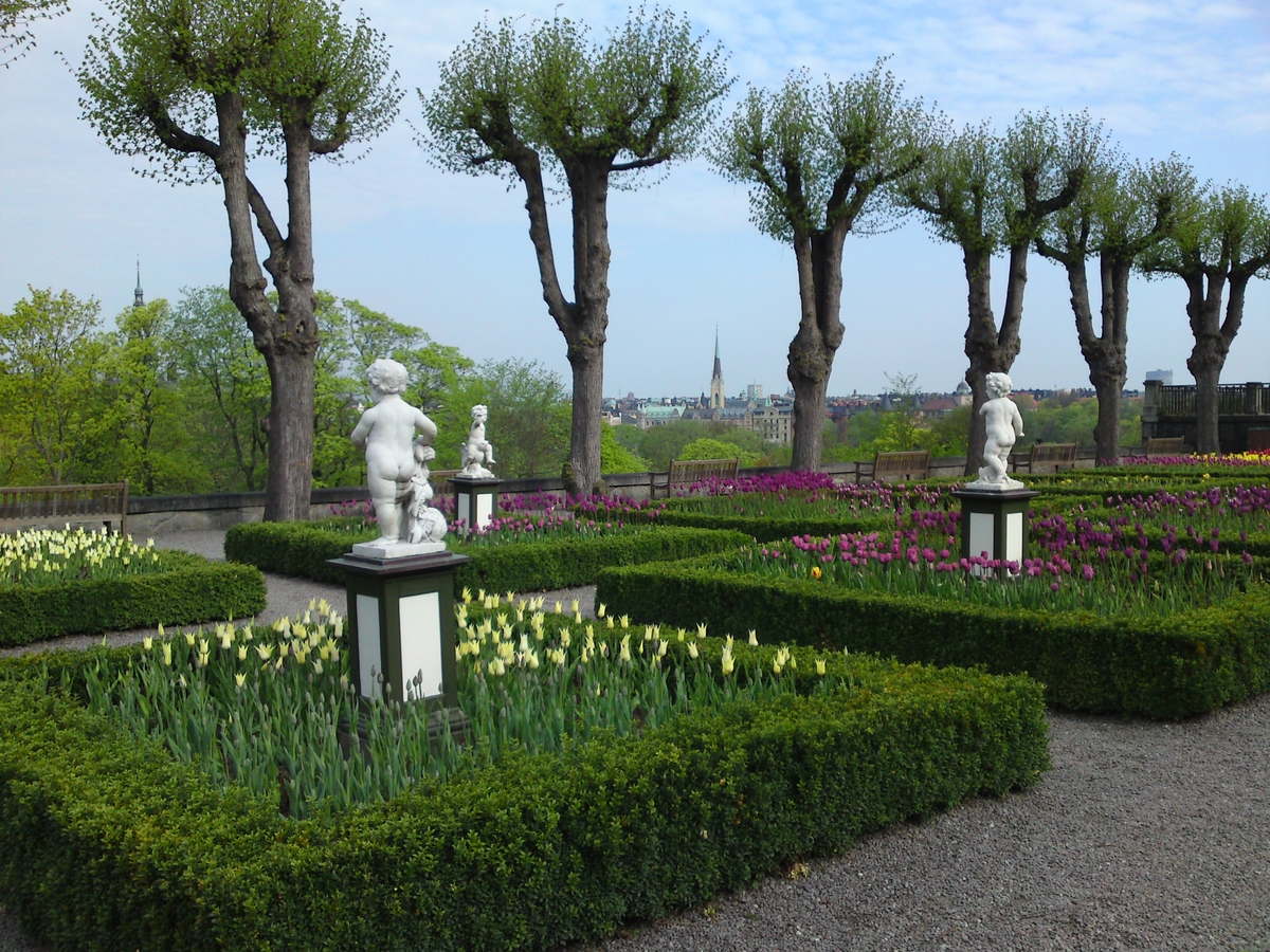 I förgrunden blommar tulpaner i olika färger övervakade av några figurer. Träd i rad rammar in Östermalm i bakgrunden.