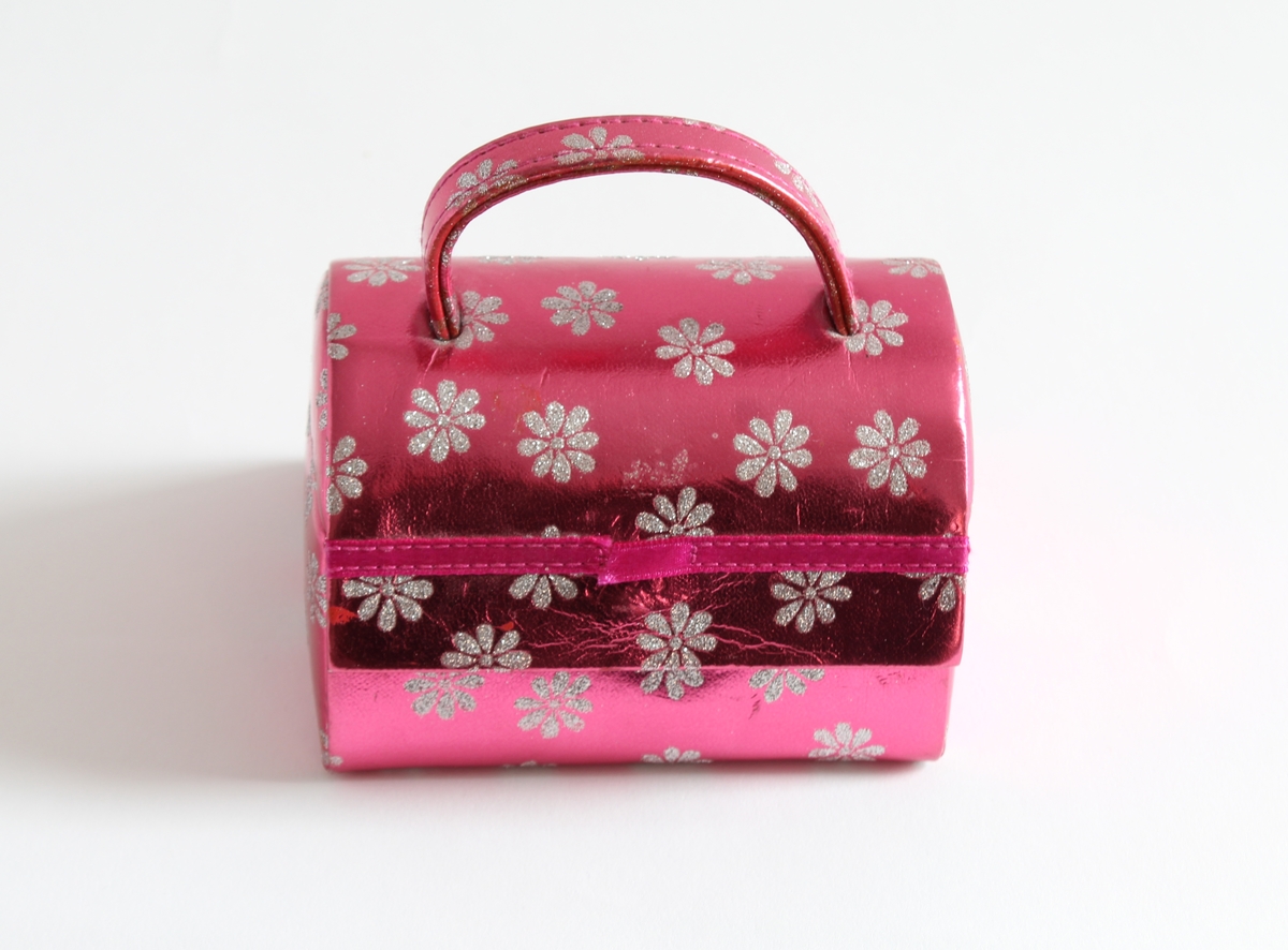 Tilhørt jente (f. 2001) i Sør-Odal. 
Et rosa bærbart smykkeskrin med hvite glitter blomster. Inne i skrinet er det speil og tre ulike rom til å ha ulike smykker i.