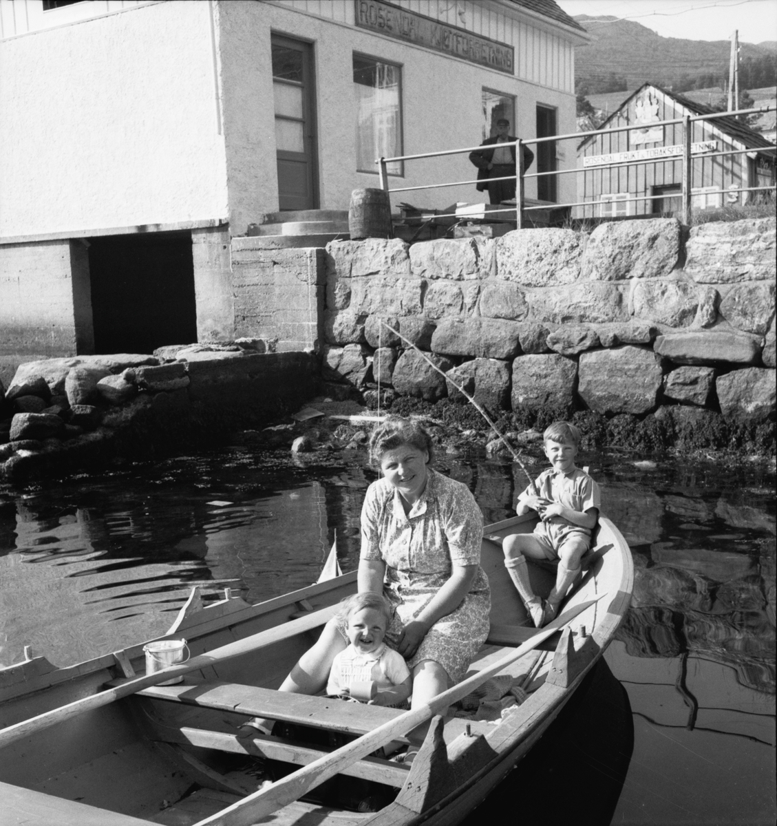 Kvinne og to barn i robåt, Rosendal kjøttforretning i bakgrunnen.