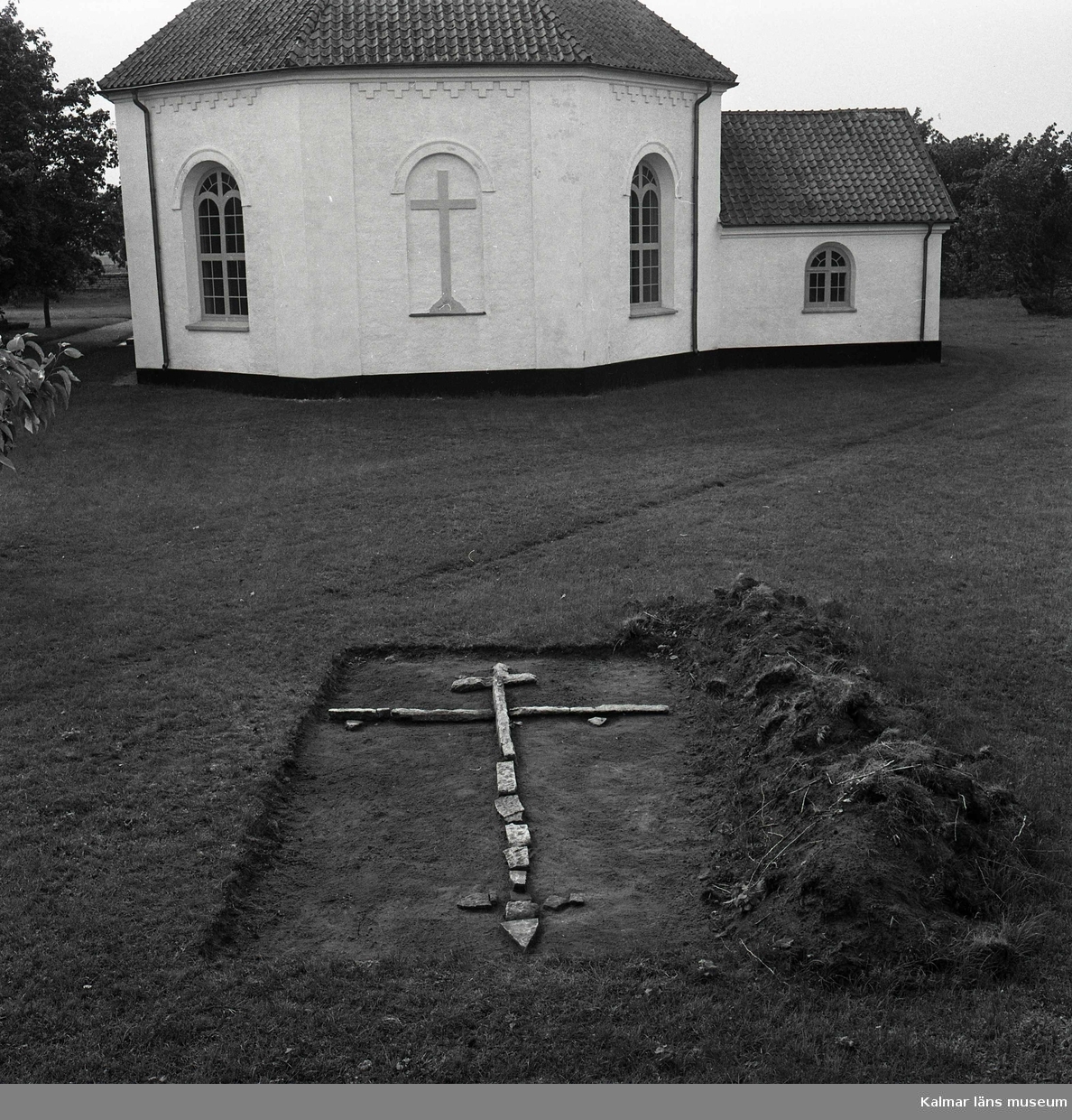 Frilagd markering av en gravplats, i form av stenar lagda i en korsformation, på Sandby kyrkogård. I bakgrunden syns kyrkan med koret närmast i bild.
