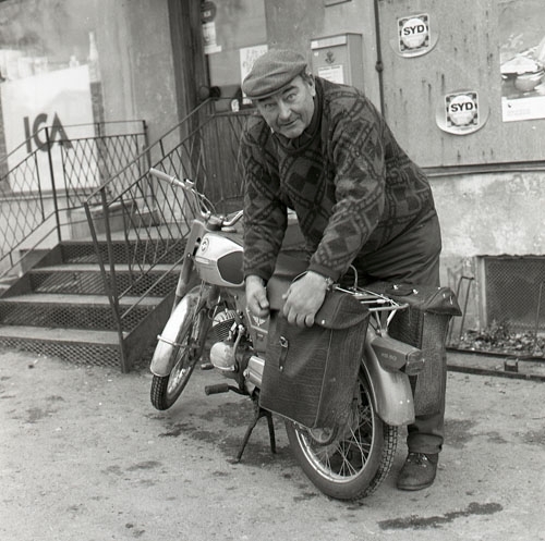Land-reportage, Brännås, Bjuråker 27 oktober 1972. En man i keps står lutad över sin moped och packar väskorna som är fästa på pakethållaren. Han befinner sig utanför en ICA-butik och vid ingången häger en postlåda. På väggen syns även reklam för läskedrycken SYD.