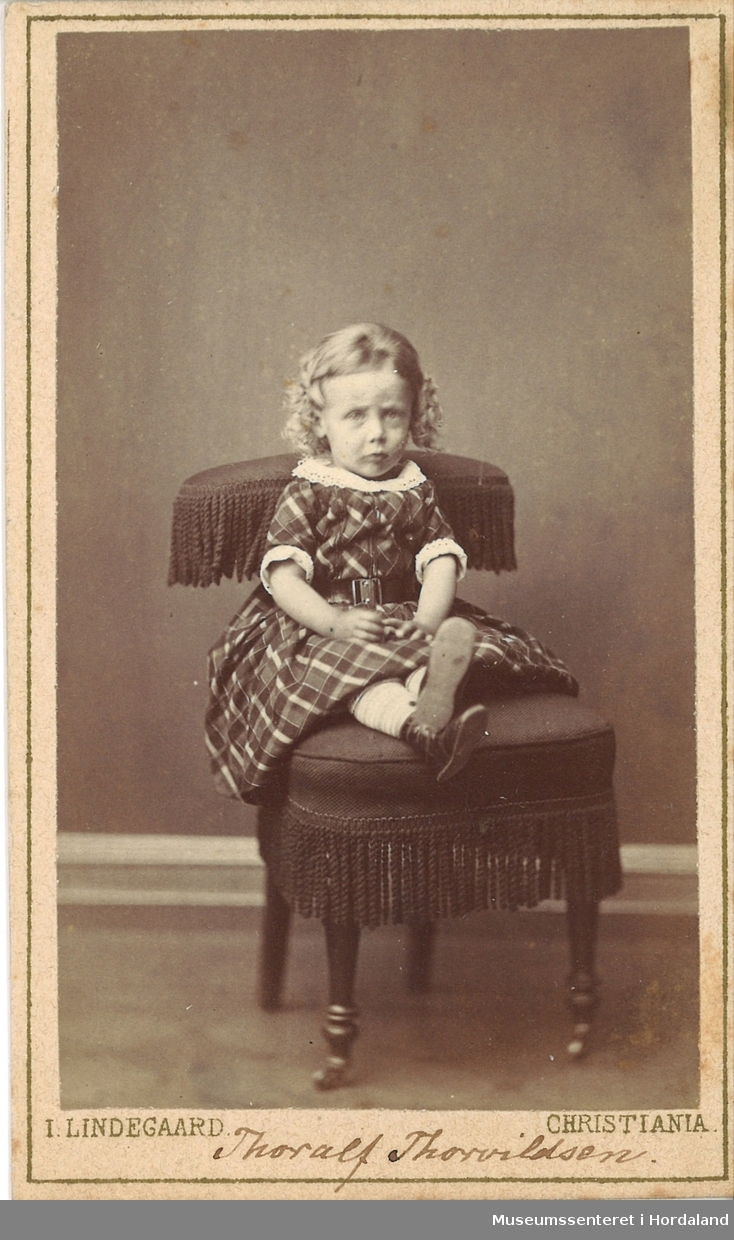 atelierfotografi av ein liten gut med ljose krøllar kledd i ruta kjole som sit på ein stol