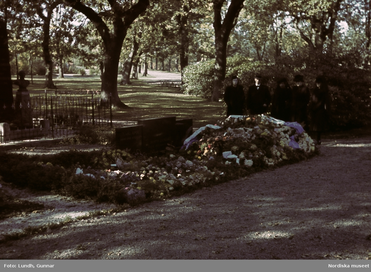 En gravplats med blommor och kistdekorationer. I bakgrunden ett sällskap begravningsklädda, fyra kvinoor och en man.