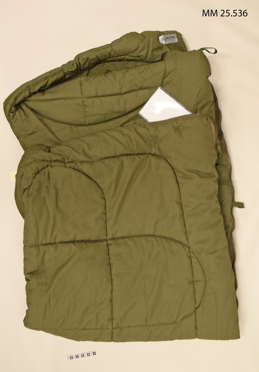 Grön sovsäck med dragkedja längs sidan och i överkant. Vidhängd lapp ger information om fastställandet.